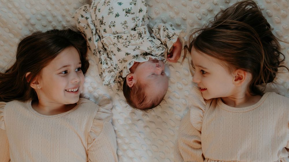 Two older sisters lying down with their newborn baby sibling between them, smiling warmly in soft neutral tones.