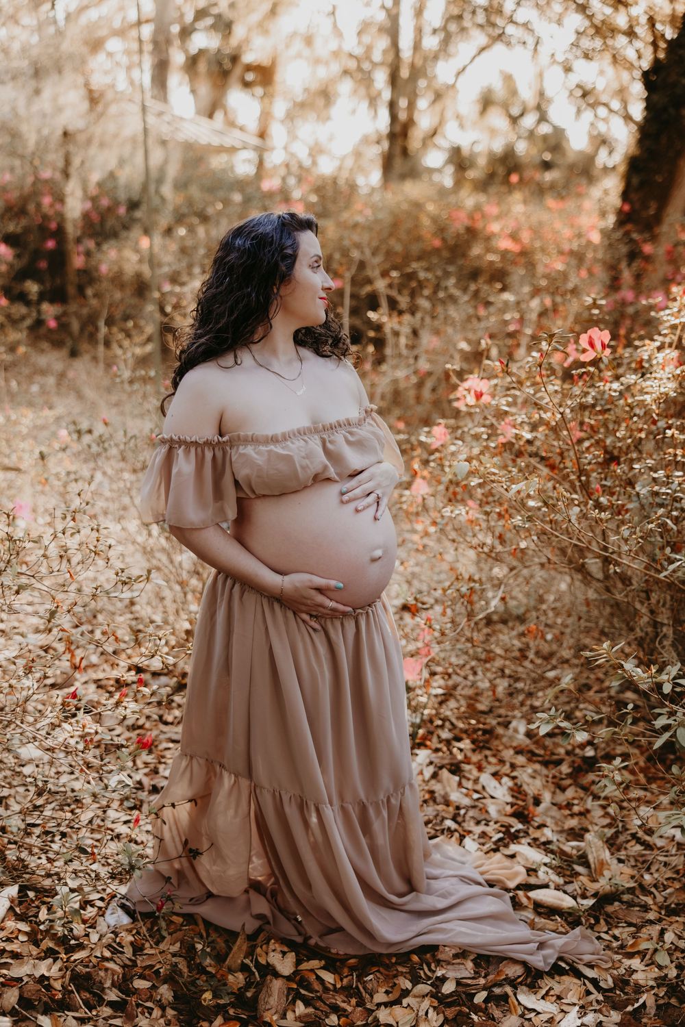 Pregnant woman in beige off-shoulder gown cradling her belly outdoors among autumn leaves and pink flowers.