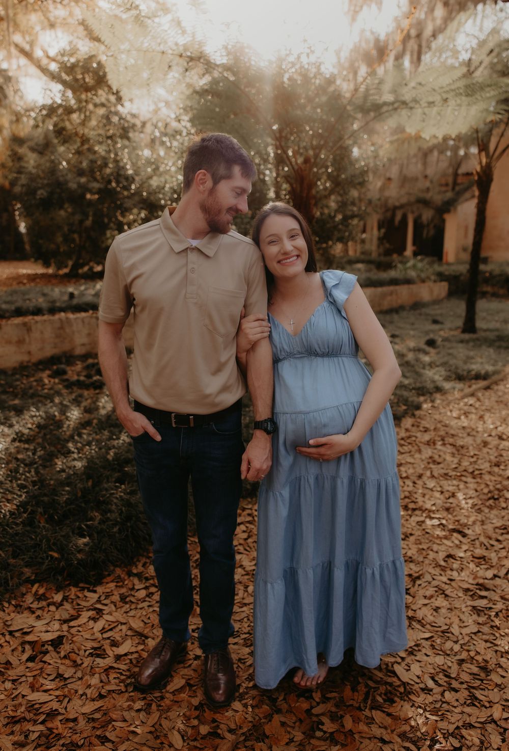 Couple poses outdoors during golden hour maternity photo shoot, woman in blue dress cradles baby bump.