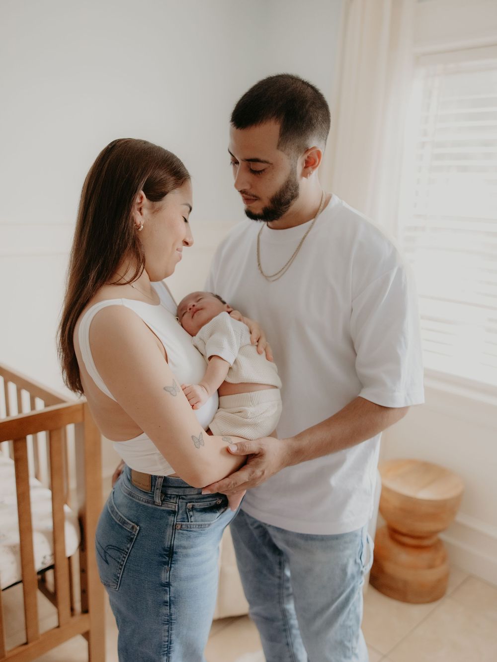 Parents in white shirts and jeans tenderly hold and gaze at their newborn baby in a bright, minimalist room.