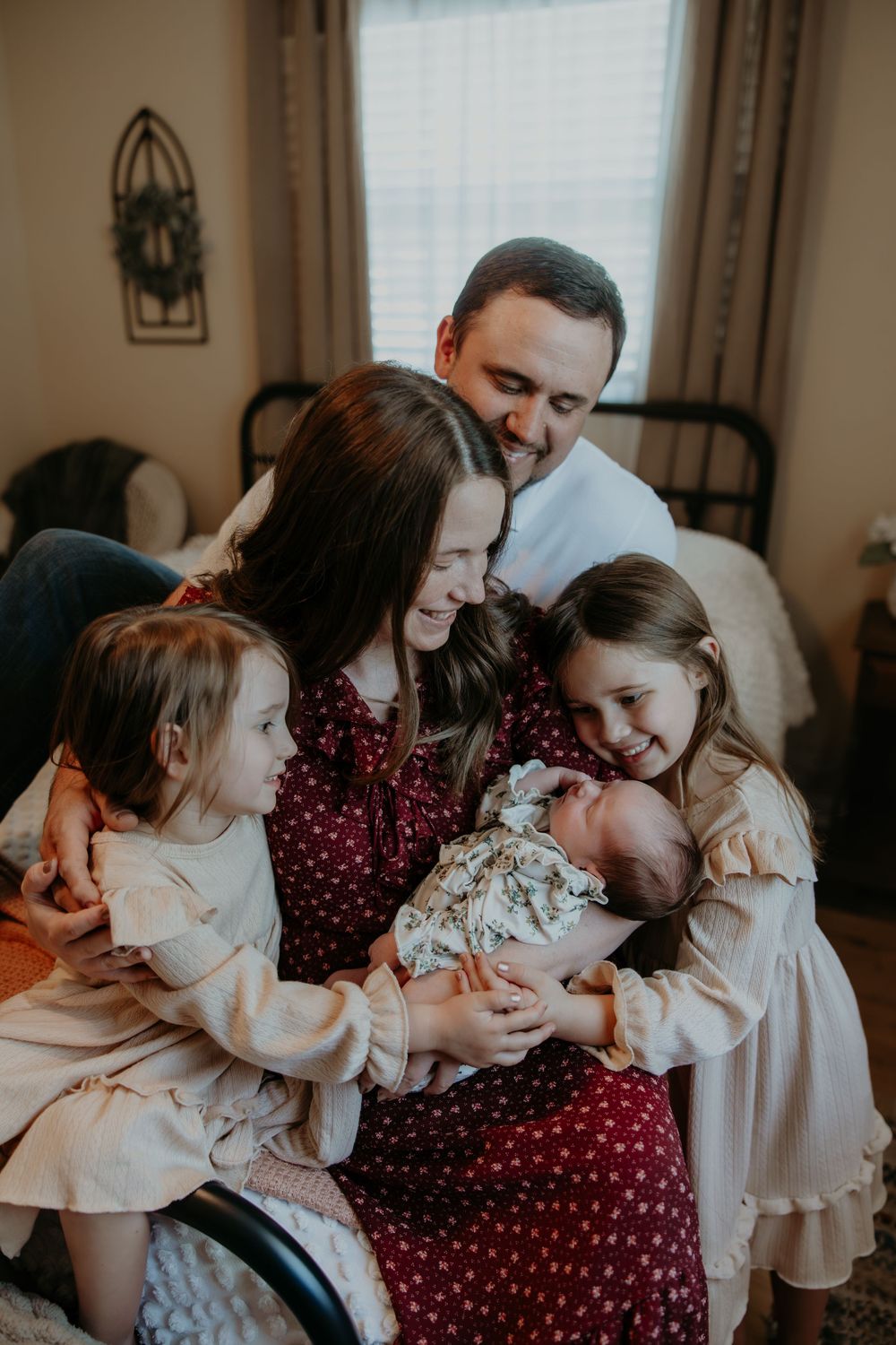 A loving family of five gathers closely on a bed, parents and two girls tenderly holding a newborn baby in a cozy bedroom.