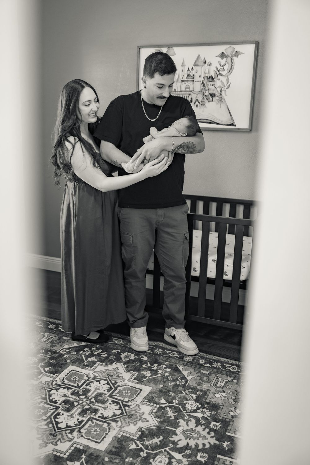 Black and white photo of new parents holding their newborn baby in a nursery with a crib and decorative rug.