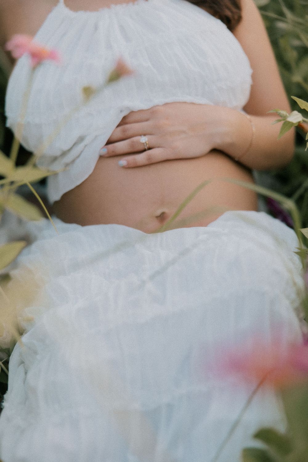 Close-up of pregnant belly in white flowing fabric with floral elements and soft pink accents in natural setting.