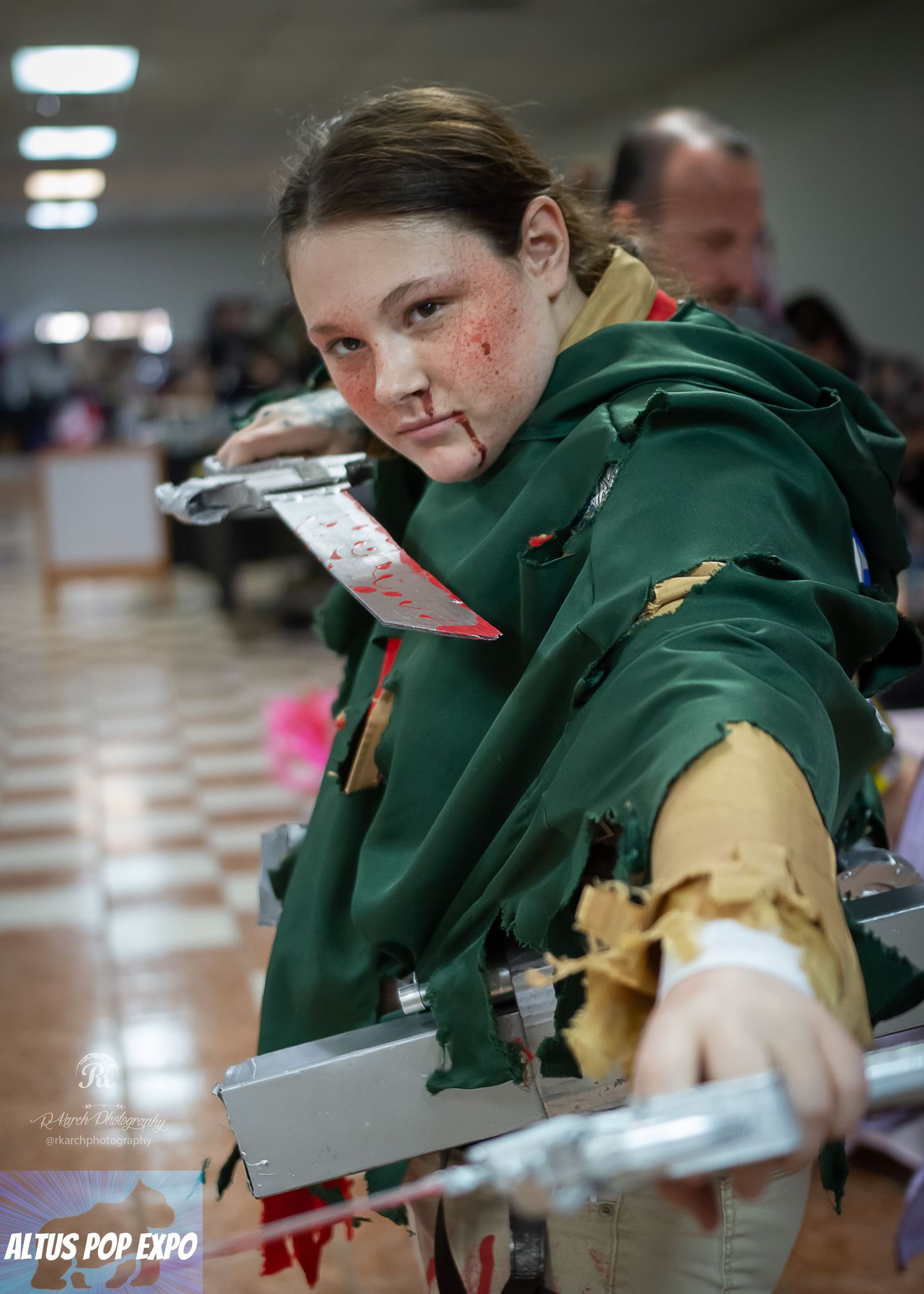 Cosplayer in green medieval-style costume holds prop weapons at convention event.