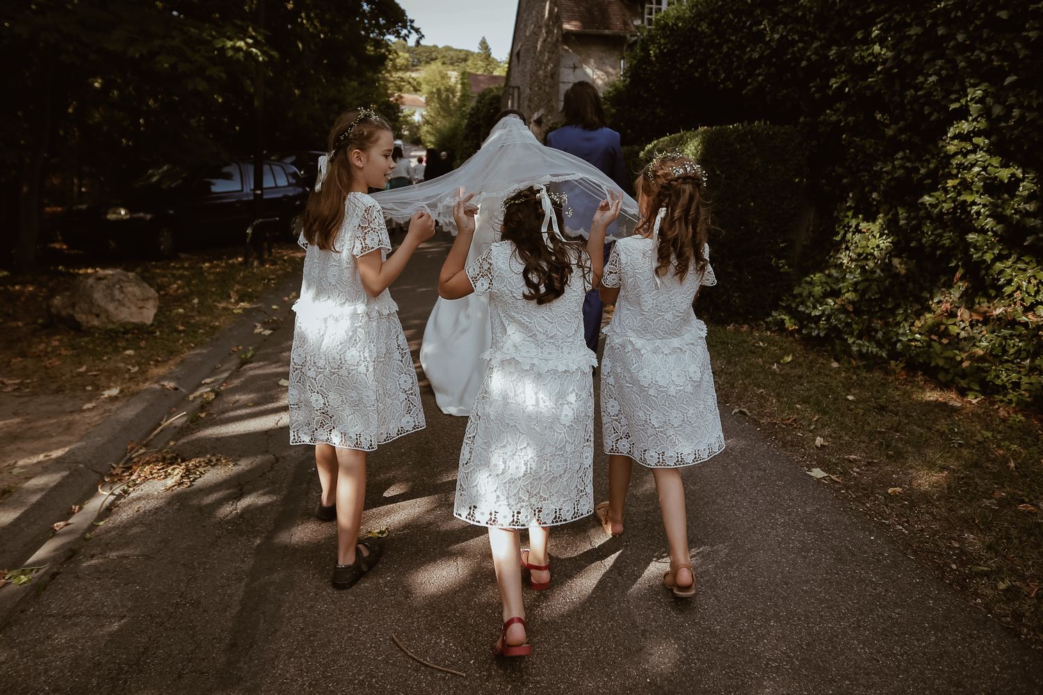 Four flower girls in white lace dresses walk down a path holding a bridal veil at an outdoor wedding.