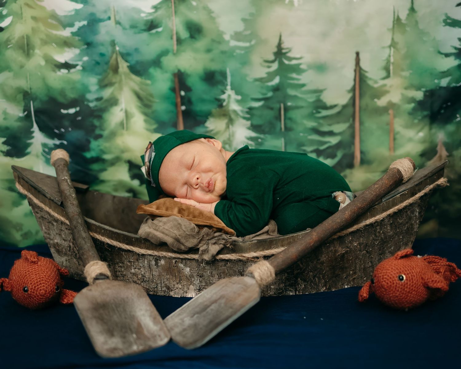 A sleeping baby in green against a painted forest backdrop with small pumpkin decorations.