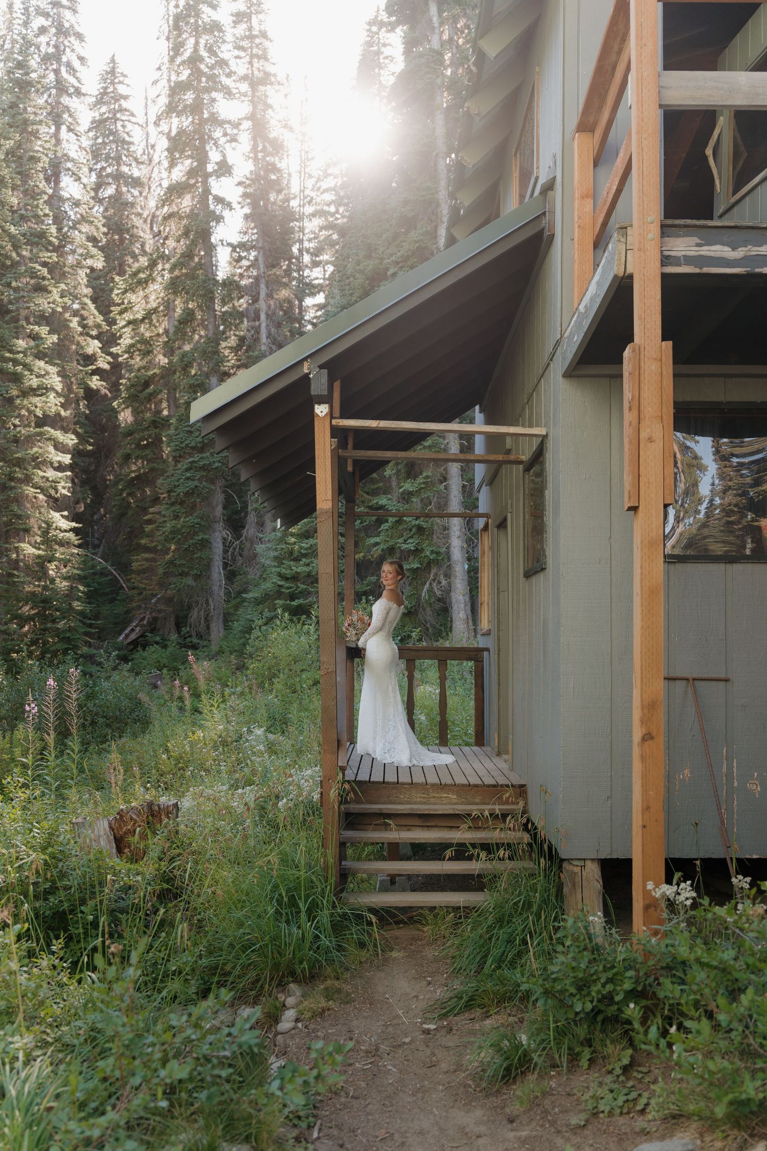A white wedding dress hangs on a rustic wooden porch railing surrounded by evergreen trees and wildflowers.