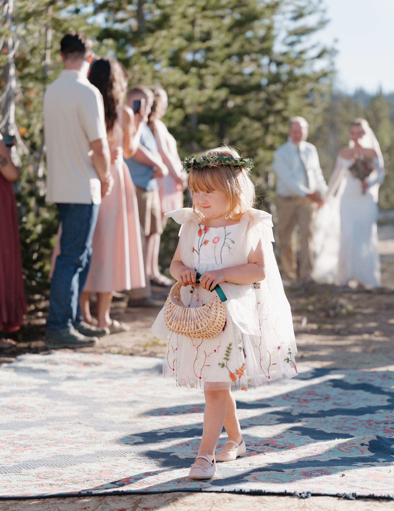 A young flower girl in a white dress and floral crown carries a wicker basket at an outdoor wedding ceremony.