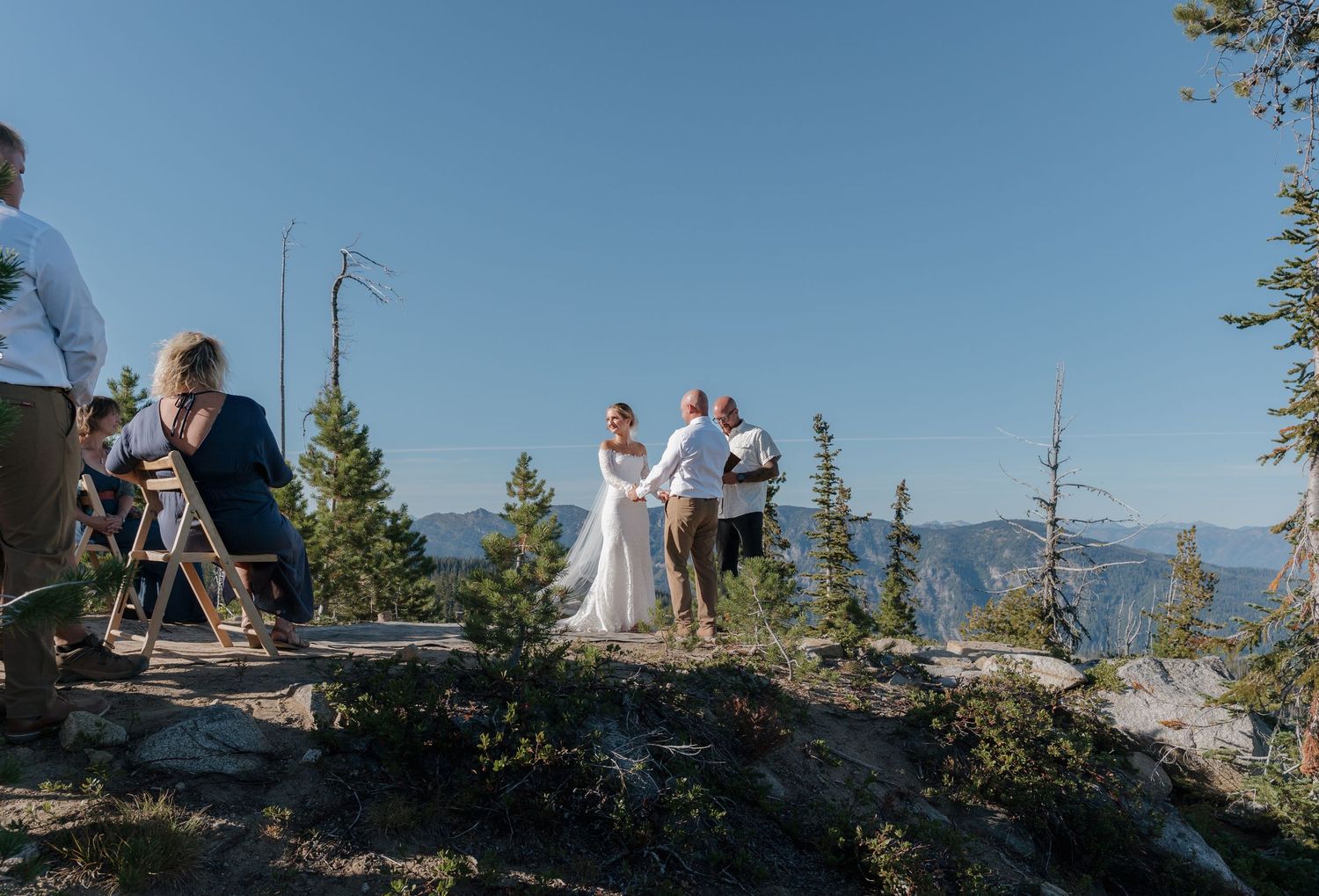 A couple poses together on a scenic mountain overlook with evergreen trees and distant peaks in view.
