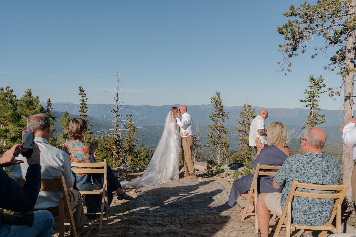 Outdoor wedding ceremony overlooking a scenic mountain lake with guests seated on wooden benches.