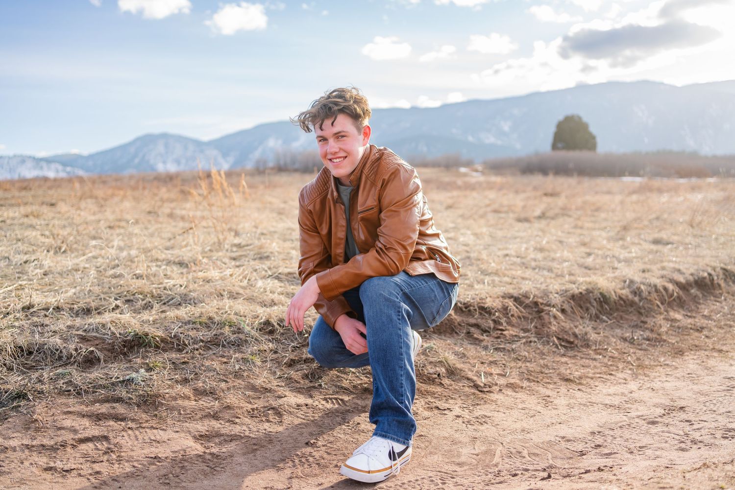 Person in brown jacket sits contemplatively on hill with mountains in background.
