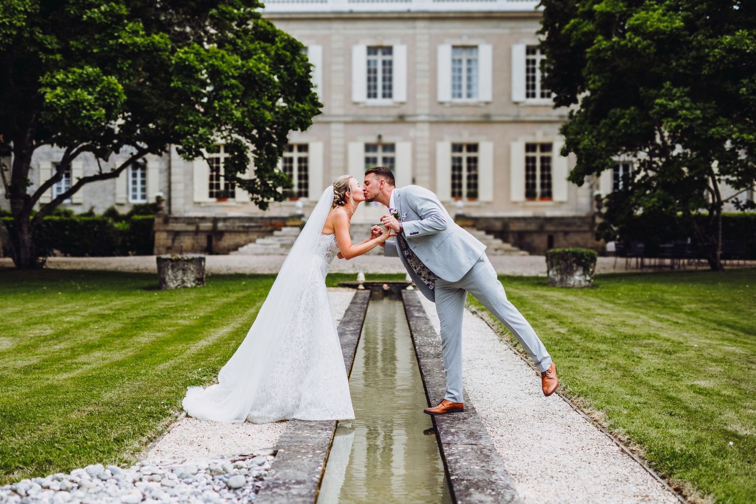 A romantic wedding photo in front of an elegant French chateau with a reflection pool and manicured lawn.