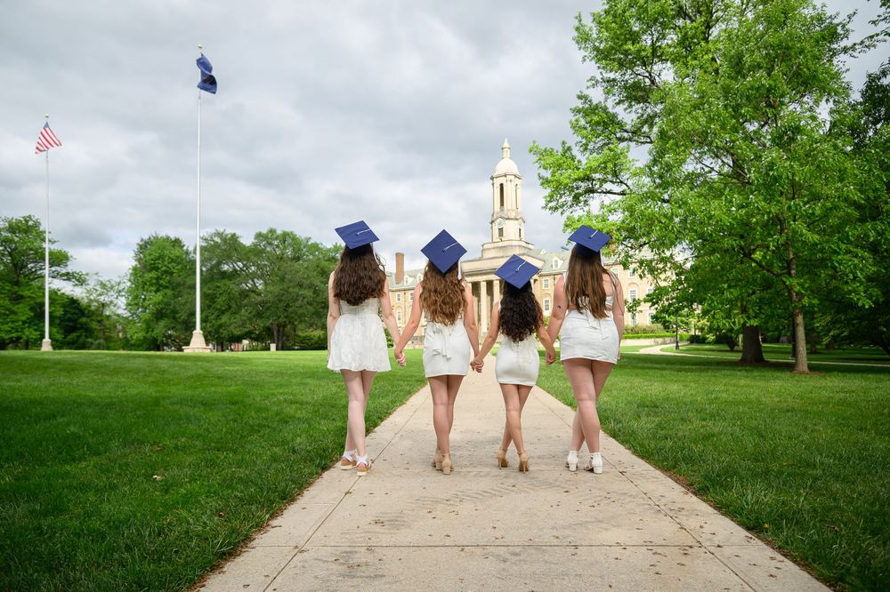 Four graduates in white dresses walk together holding their blue caps on a grassy hill during commencement.
