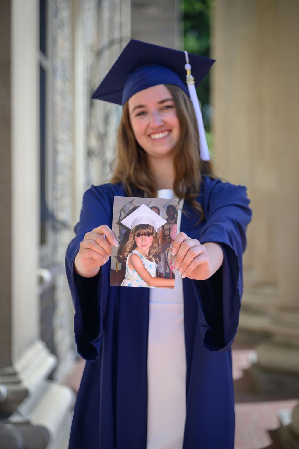 Graduate in blue cap and gown holds up and tears photo during graduation photo session at university campus.