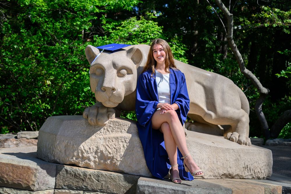 A person in a blue dress sits on a stone sculpture in a garden setting with lush greenery in the background.