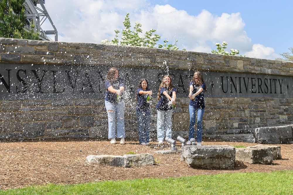 Students sit along an old stone wall on a sunny day at a university campus.