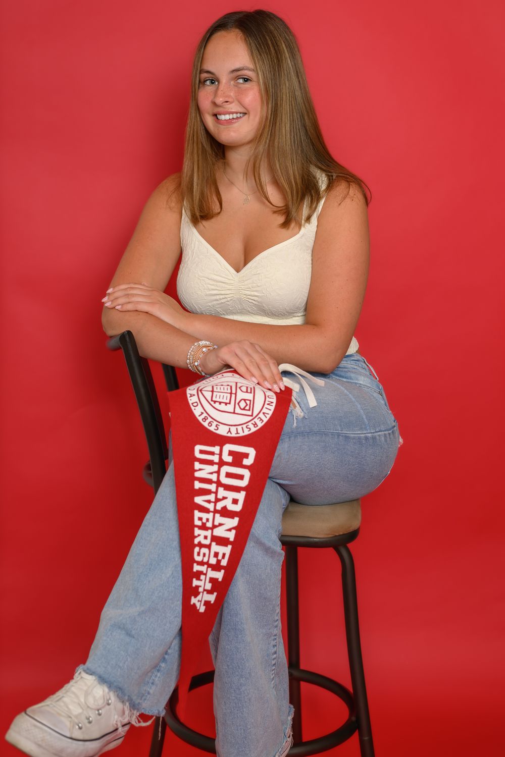 A person in jeans and a white top poses casually on a black chair against a bright red background.