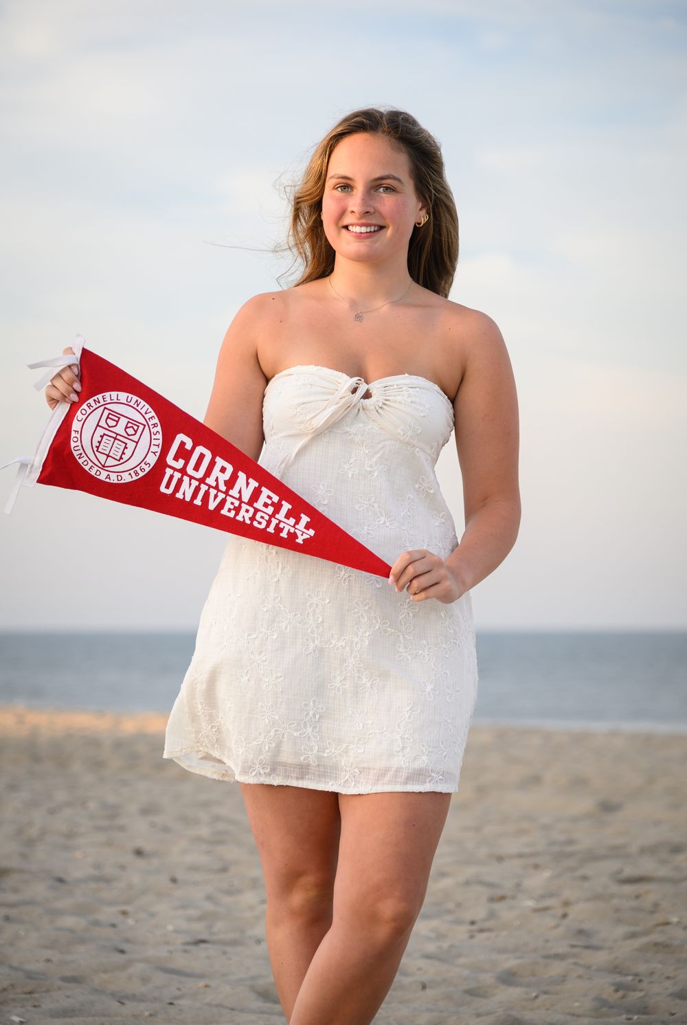 Someone in a white lace dress holds a red Cornell University pennant while standing on a beach at sunset.