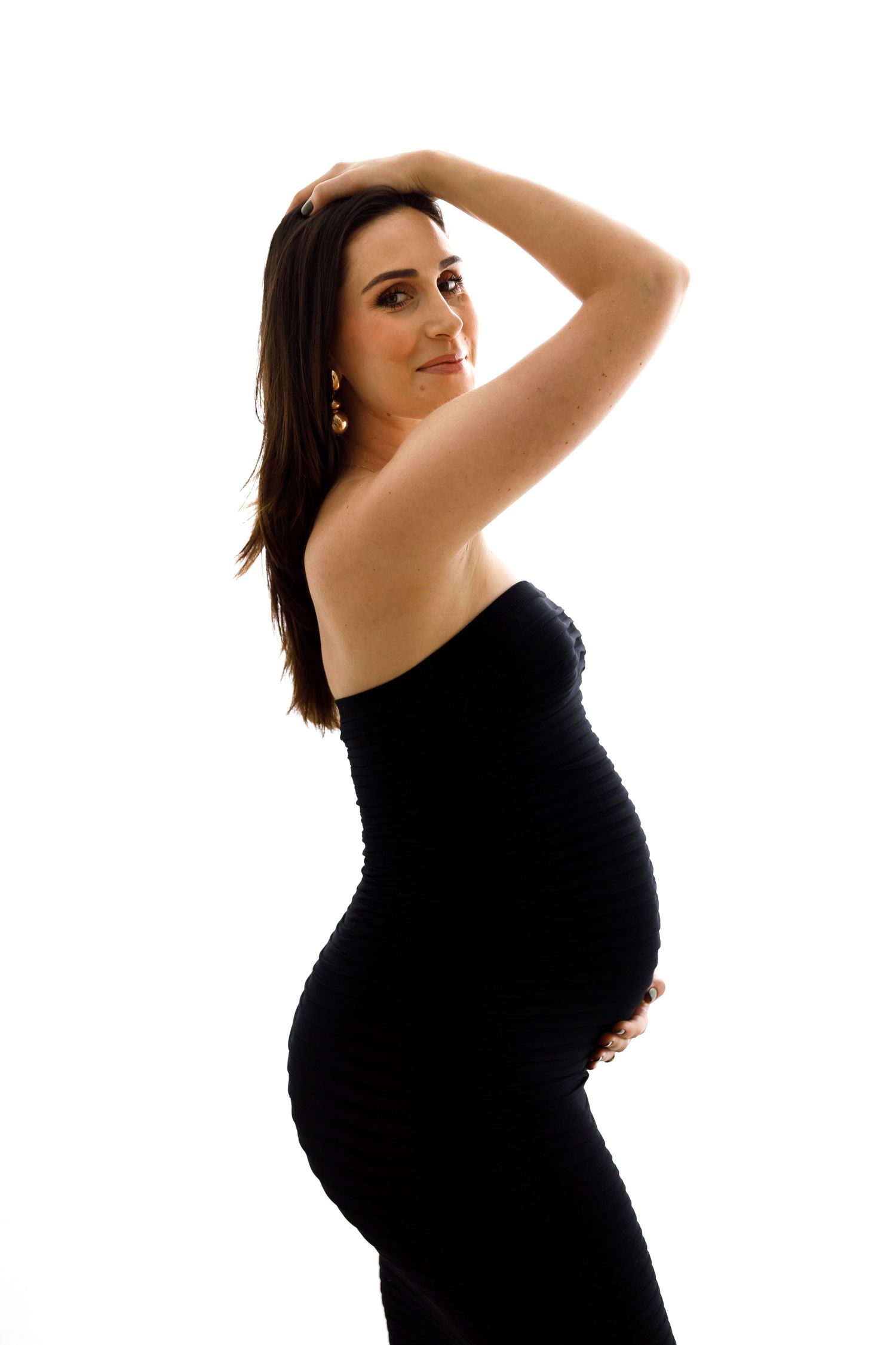Professional maternity portrait of an expectant mother in a black dress posing against white background.