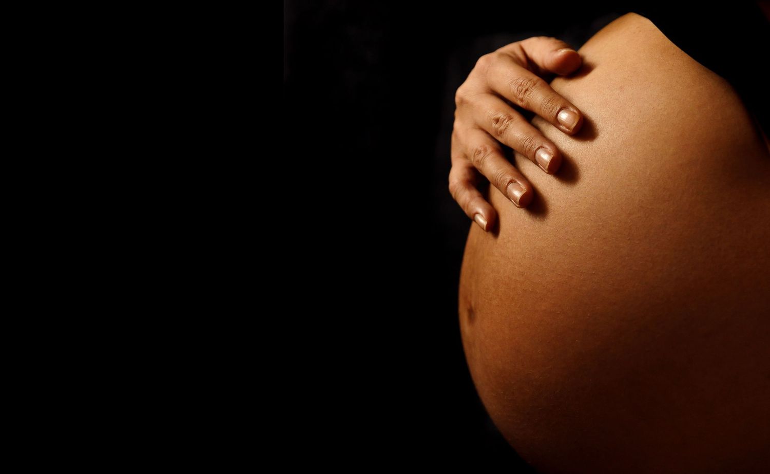 Close-up silhouette of a pregnant belly with hands gently resting on top against dark background.