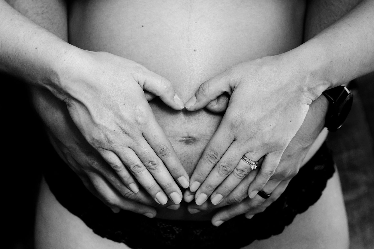 Black and white close-up of hands forming heart shape on pregnant belly.