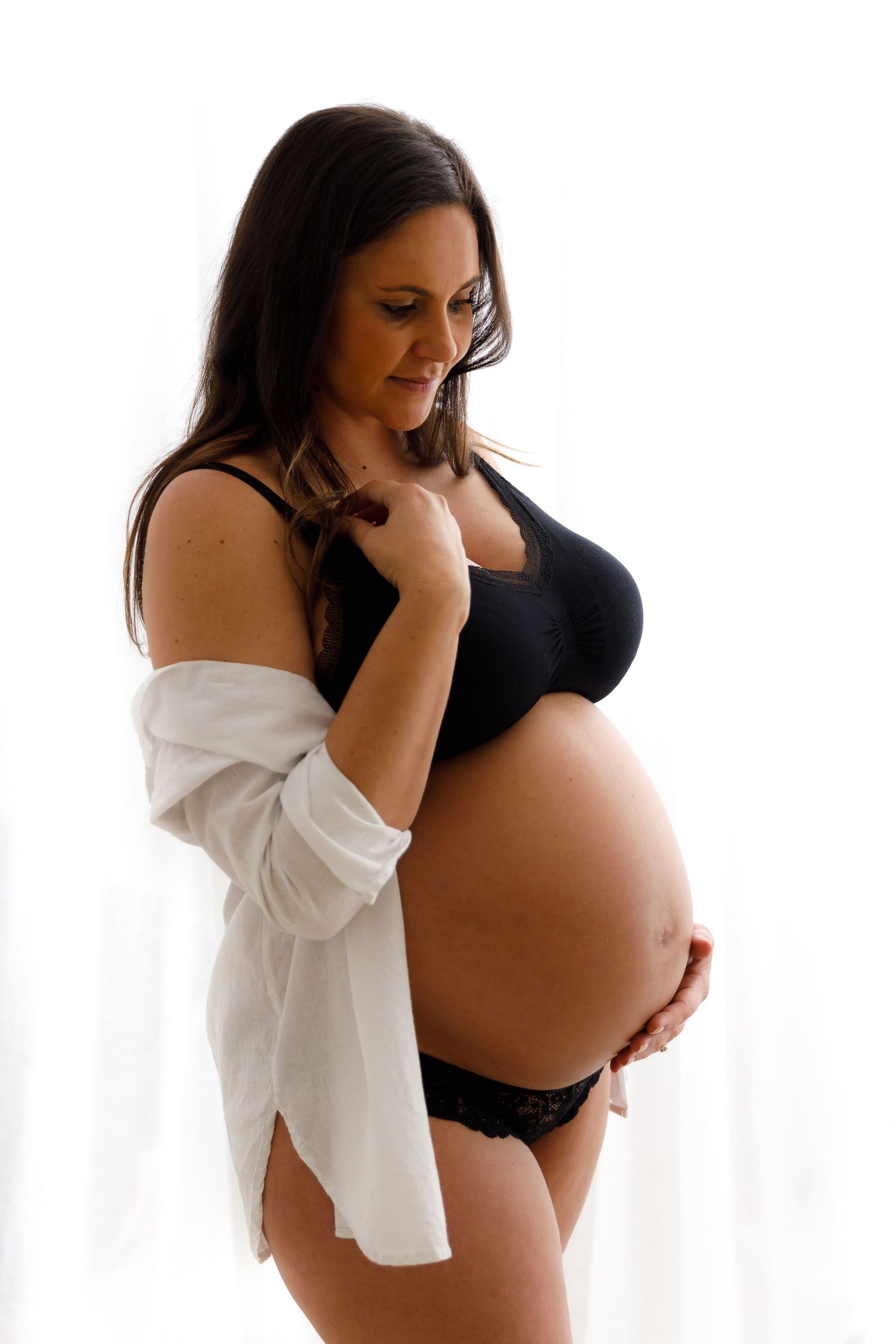Side view of pregnant woman in intimate apparel and white shirt against white backdrop.
