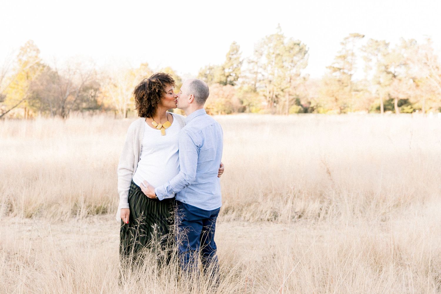 Romantic maternity photo of couple standing together in sunlit field.