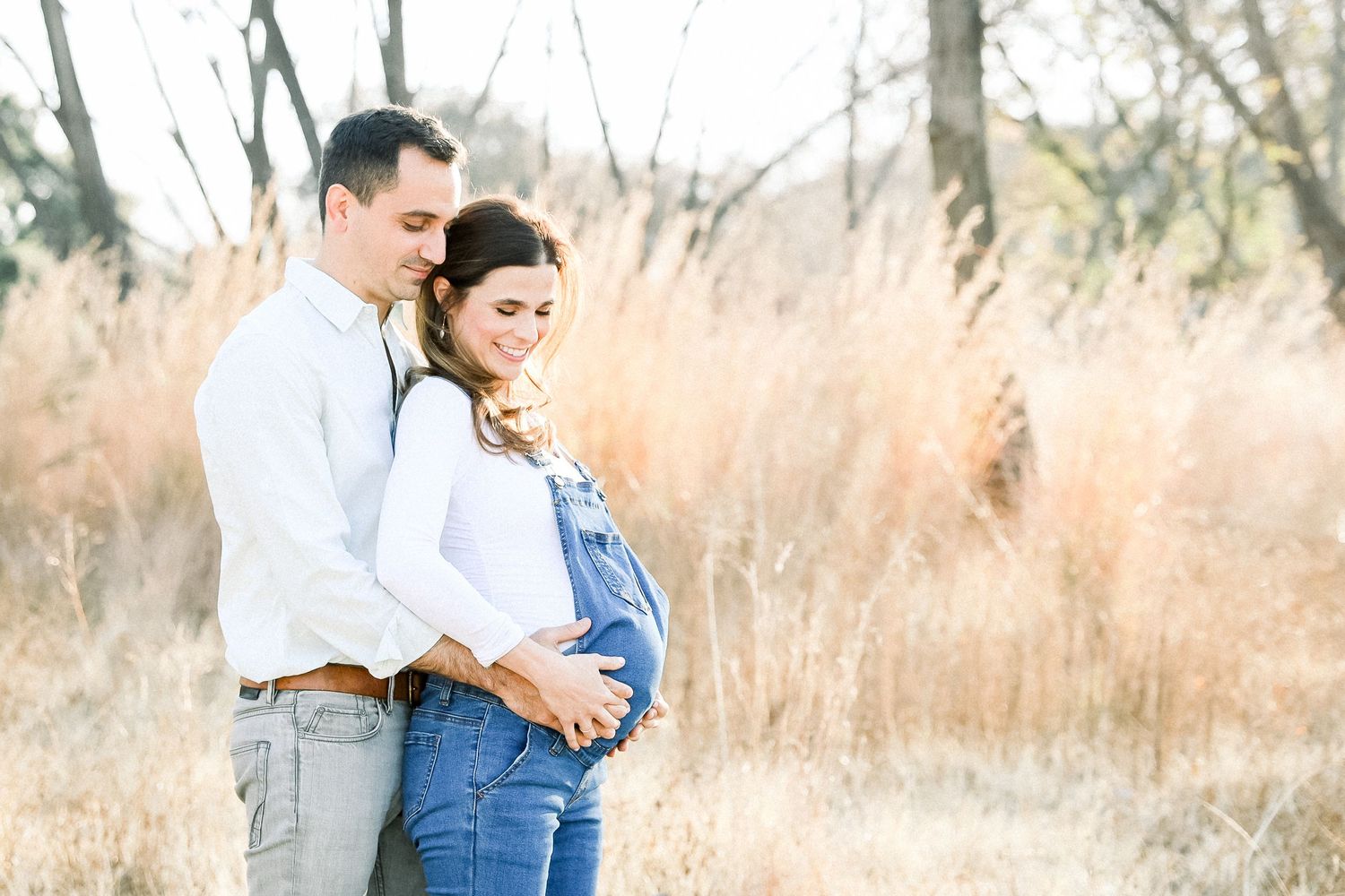 A couple in casual attire embracing in a sunlit field with tall grass during a maternity photoshoot.