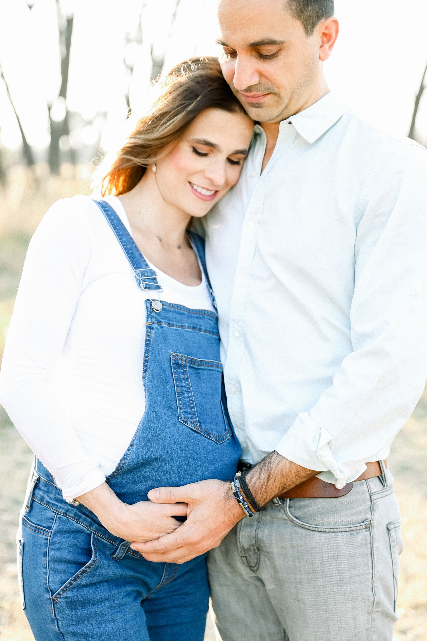 A tender maternity moment captured in overalls against a natural outdoor backdrop with soft lighting.
