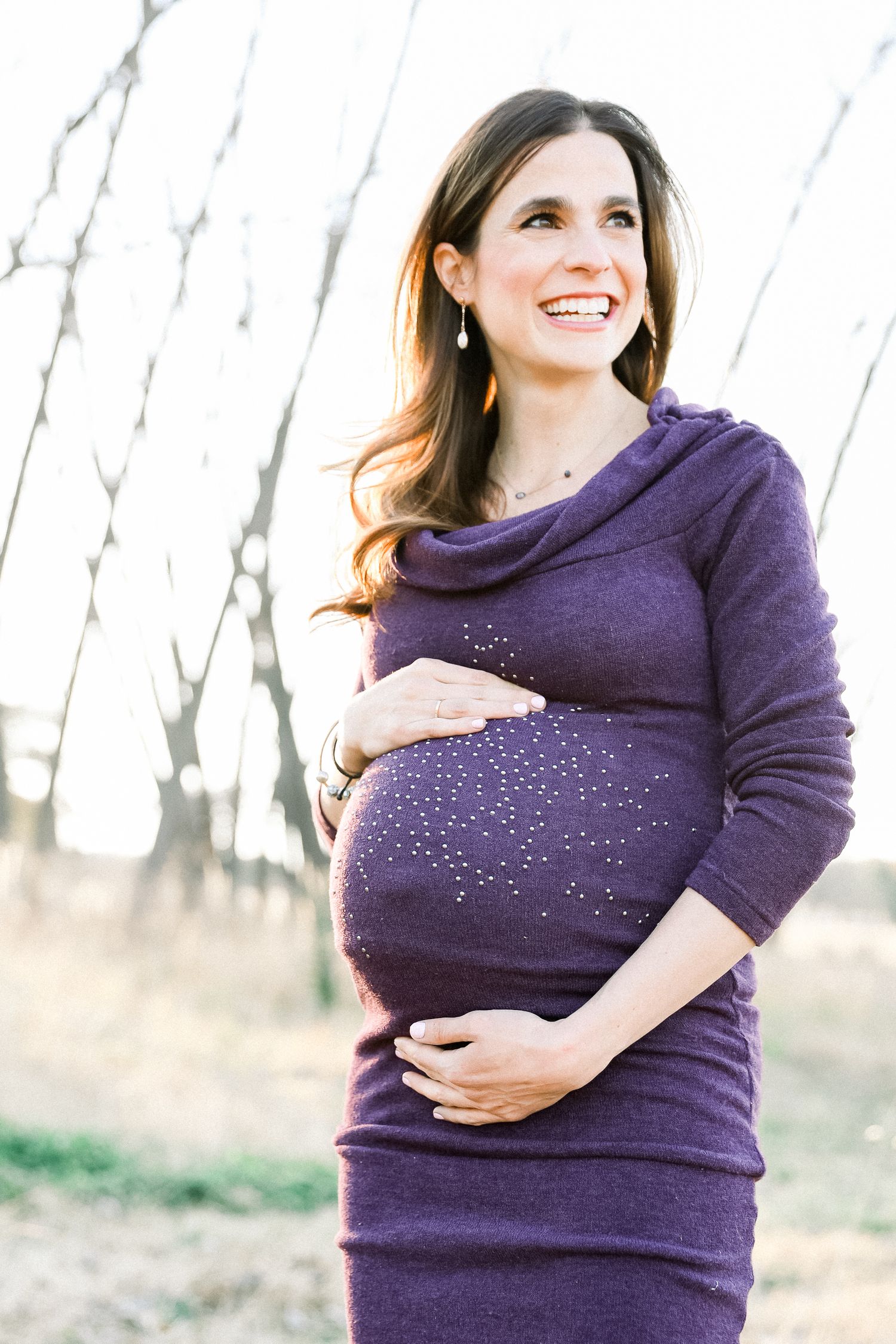 An expectant mother in a purple sparkly sweater dress smiling outdoors during winter.