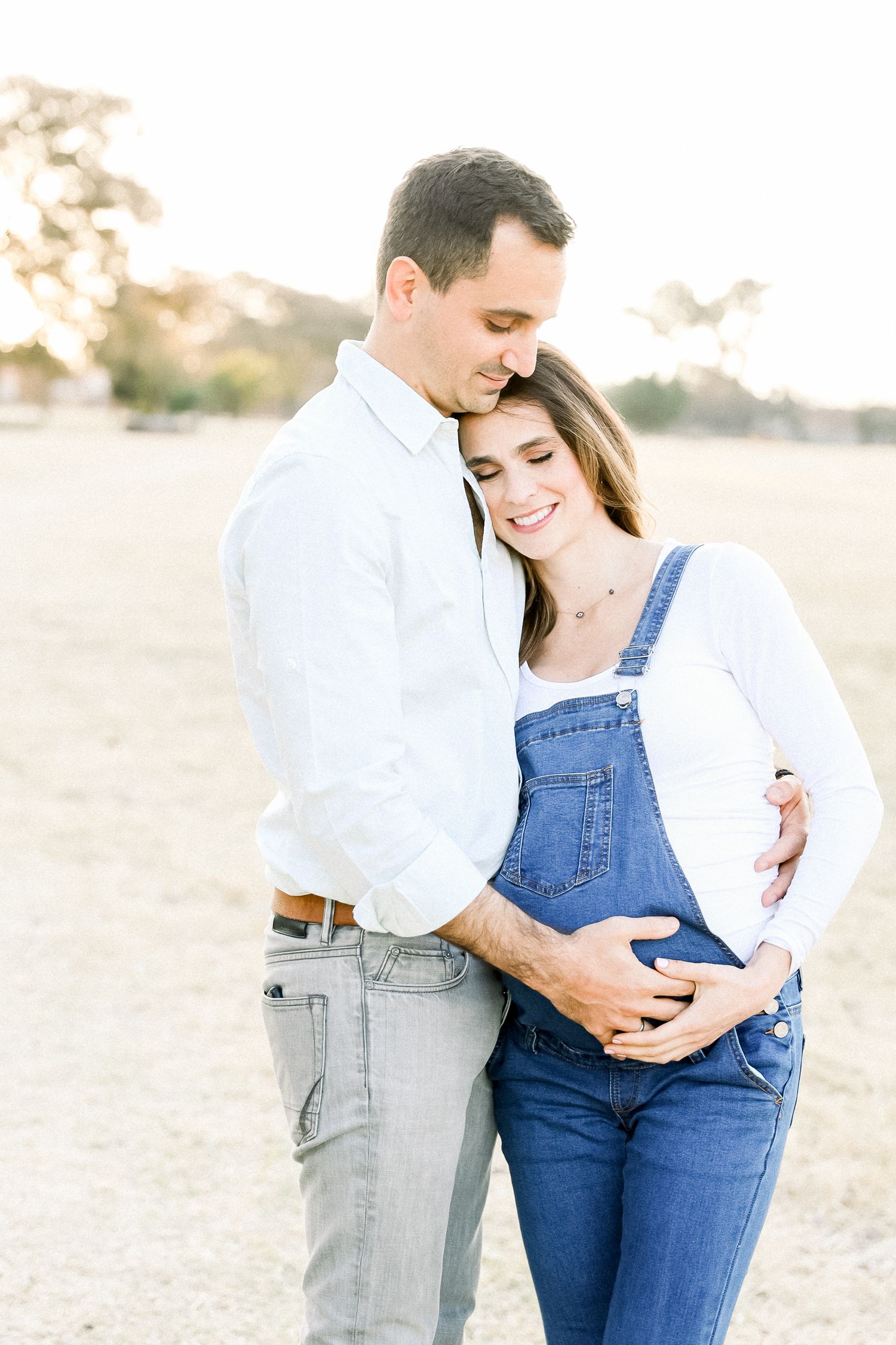 A maternity photoshoot in denim overalls and white shirt against a warm sunset background.