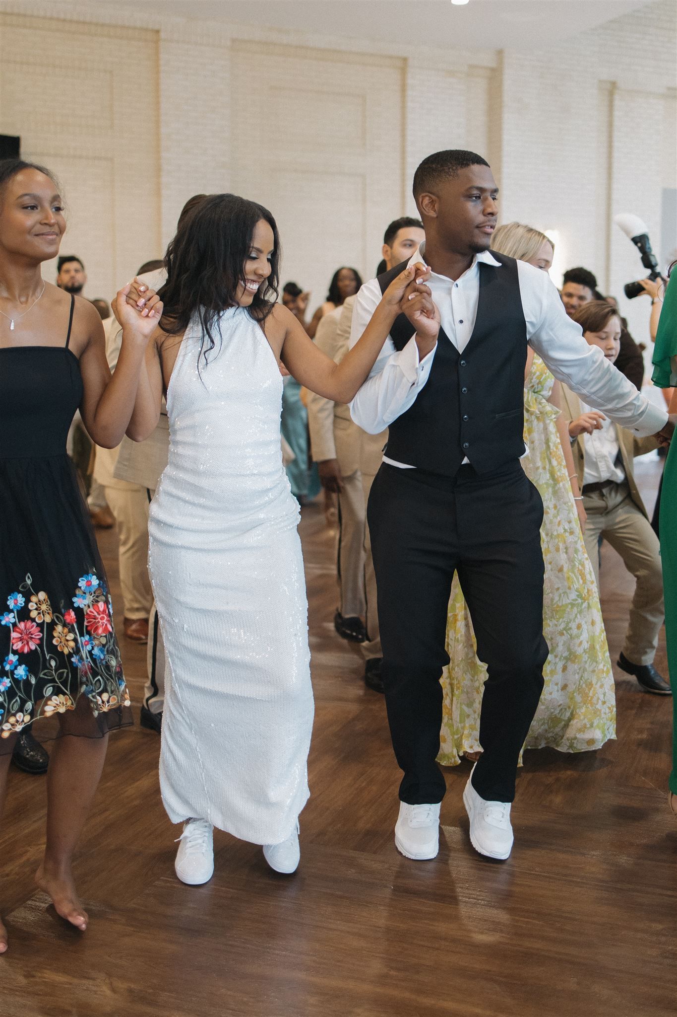 Wedding guests dancing together on hardwood floor during reception with woman in white dress and man in black vest.