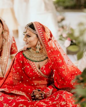 Traditional bride in red embroidered lehenga with ornate gold jewelry at outdoor ceremony.