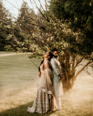 Artistic portrait of couple in South Asian attire posing against tree in golden sunlight.