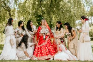 Bridal party in ivory gowns gathers around bride wearing stunning red Indian wedding attire under willow branches.
