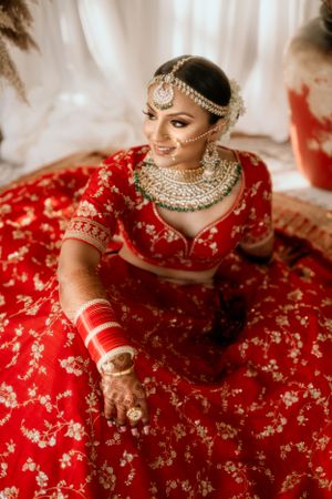 Bride in red wedding lehenga with intricate gold jewelry poses against white backdrop.