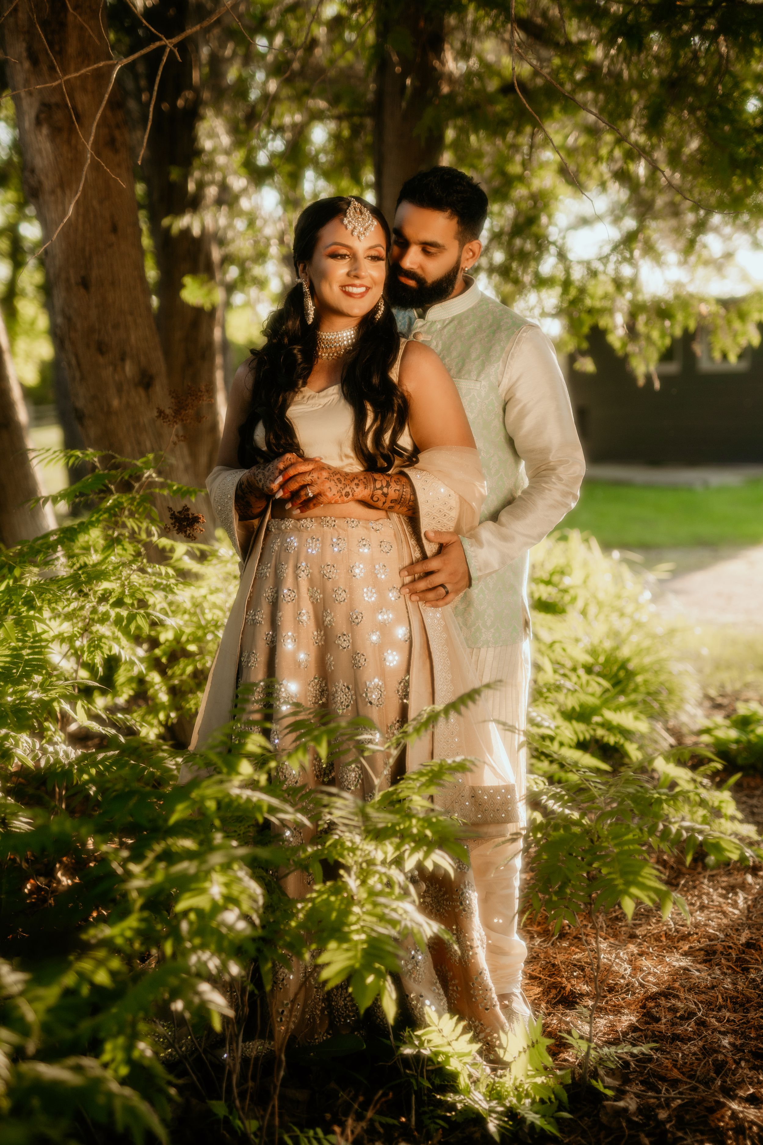 Couple in traditional South Asian attire share an intimate moment in a sunlit garden.