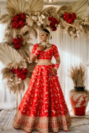 Traditional red wedding lehenga photographed against dried pampas grass and red floral backdrop.