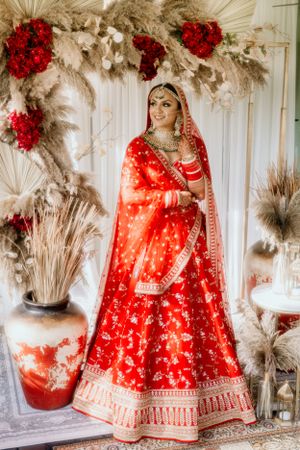 Elegant red bridal outfit with gold embellishments photographed under dried pampas grass decor.