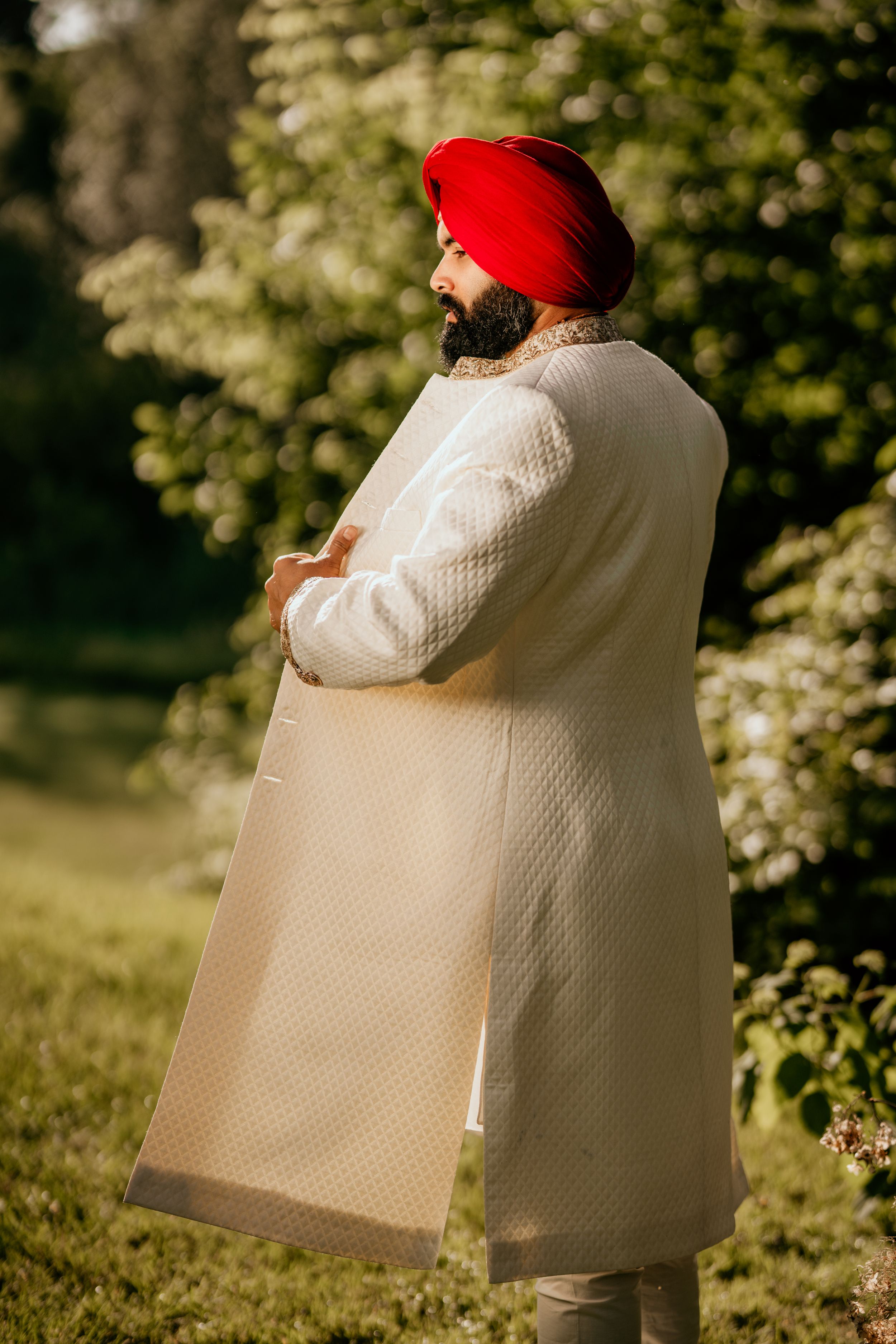 Groom wearing a traditional white sherwani and red turban poses in a garden setting during an Indian wedding.