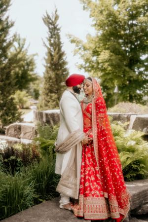 Traditional Sikh wedding couple in red and white attire share an intimate moment in a garden setting.