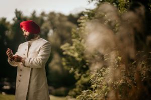 Groom in cream sherwani and red turban adjusts his accessory in an outdoor garden setting.