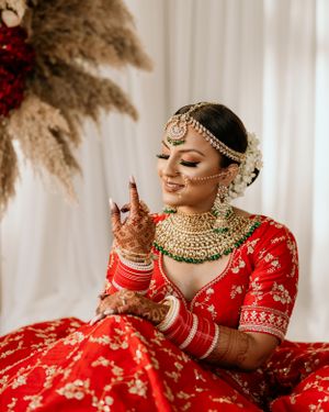 Traditional bride in red lehenga with ornate gold jewelry and henna sits against white backdrop with pampas grass.