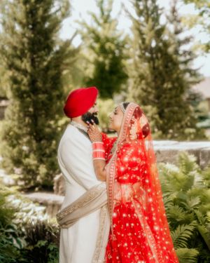 A couple in traditional Sikh wedding attire embraces against a backdrop of evergreen trees.