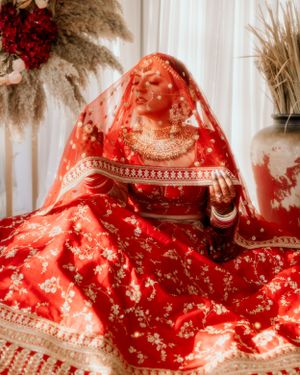 Bride in red bridal lehenga with veil and jewelry sits against white backdrop with pampas grass decor.
