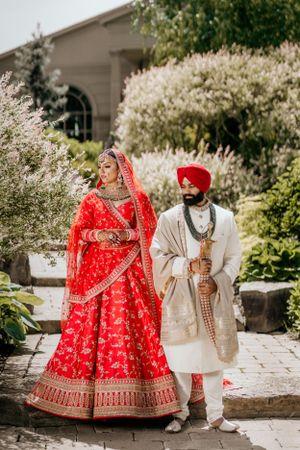 Sikh bride in red lehenga and groom in white sherwani pose in a garden with blooming flowers.