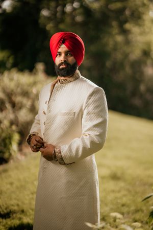 Groom in elegant white sherwani and red turban poses outdoors against natural greenery.