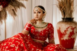 Bride in red embroidered lehenga with traditional jewelry poses against white backdrop with dried pampas grass.