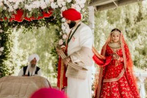 Traditional Sikh wedding ceremony with couple in red and white attire under floral canopy.
