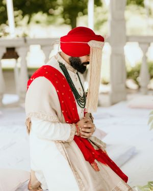 Traditional Sikh groom in white attire and red turban holding ceremonial sword.